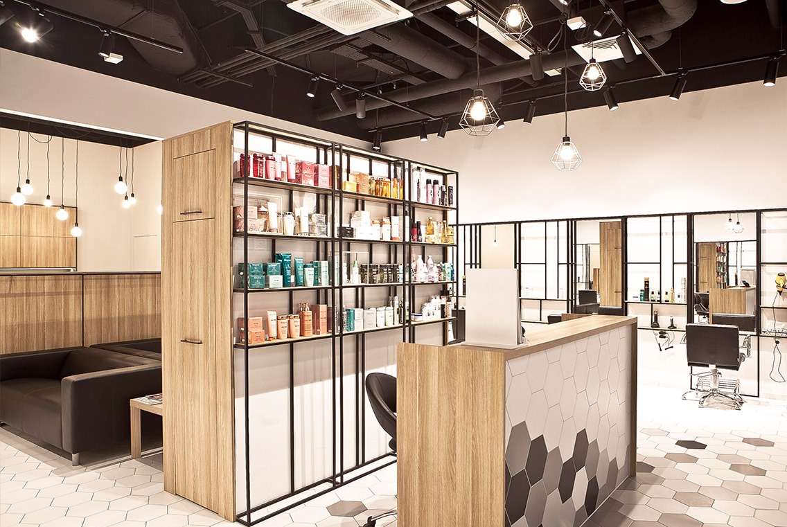 Projekt salonu fryzjerskiego: recepcja z heksagonalnymi płytkami w odcieniach szarości i drewna, regał z produktami kosmetycznymi, czarna sofa i designerskie lampy wiszące.