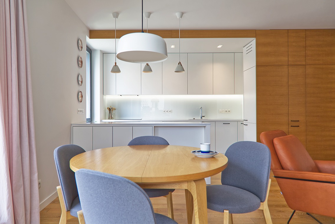 Jasne, nowoczesne wnętrze mieszkania: okrągły drewniany stół z czterema szarymi krzesłami, biała kuchnia w tle, minimalistyczne oświetlenie sufitowe i akcenty z drewna.