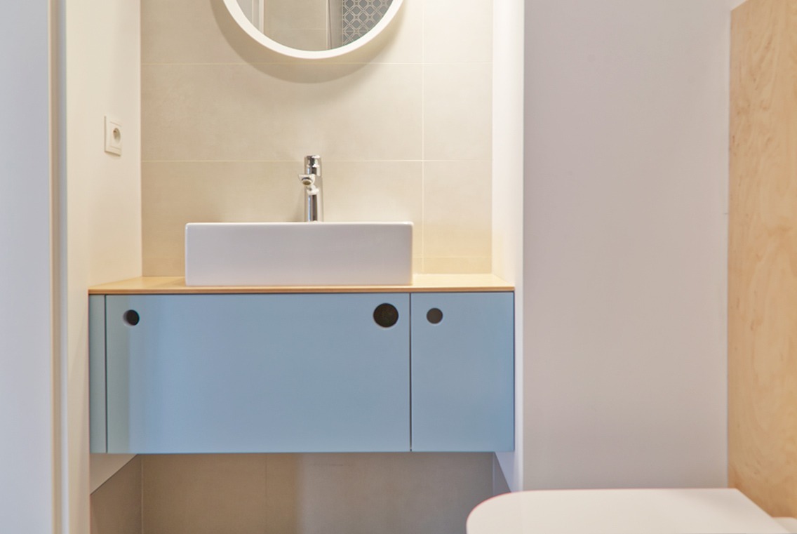 Nowoczesna łazienka z białą, prostokątną umywalką nablatową, okrągłym lustrem i szafką pod umywalką w kolorze pastelowego błękitu z okrągłymi uchwytami.