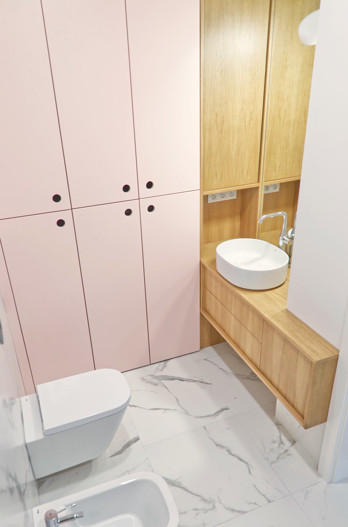 Nowoczesna łazienka z różowymi szafkami, drewnianym blatem z owalną umywalką, podwieszanym WC i bidetem na marmurowej posadzce.