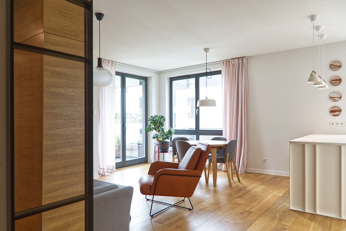 Jasne wnętrze mieszkania w Gdyni z drewnianą podłogą, oknami balkonowymi z jasnoróżowymi zasłonami, okrągłym stołem z szarymi krzesłami, brązowym fotelem i dekoracyjnymi lampami.