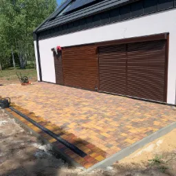 Żółto-brązowa kostka brukowa ułożona przed budynkiem z brązową bramą garażową, widoczny drenaż liniowy, częściowo zasypana piaskiem obrzeża kostki, w tle taczka i zagęszczarka.