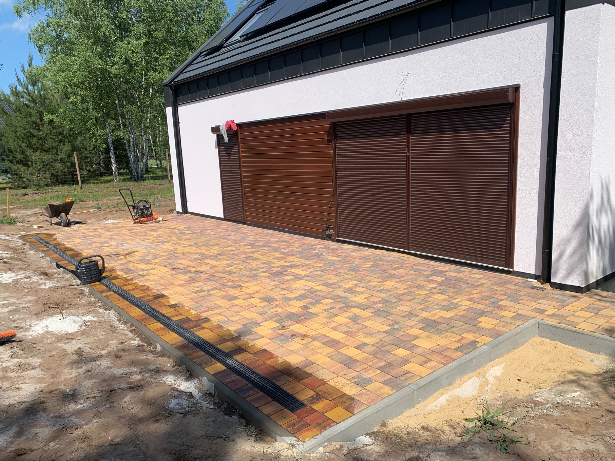 Żółto-brązowa kostka brukowa ułożona przed budynkiem z brązową bramą garażową, widoczny drenaż liniowy, częściowo zasypana piaskiem obrzeża kostki, w tle taczka i zagęszczarka.