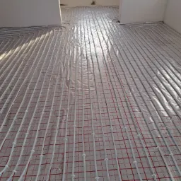 Białe rury ogrzewania podłogowego ułożone na folii z czerwoną siatką w surowym wnętrzu, przygotowane do zalania wylewką.