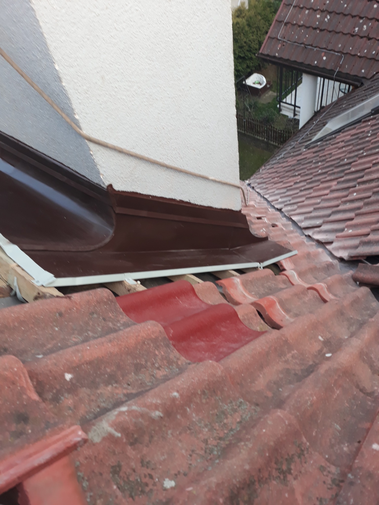 Fragment dachu z czerwonej dachówki ceramicznej, widoczna nowa, brązowa obróbka blacharska komina i fragment konstrukcji więźby dachowej.