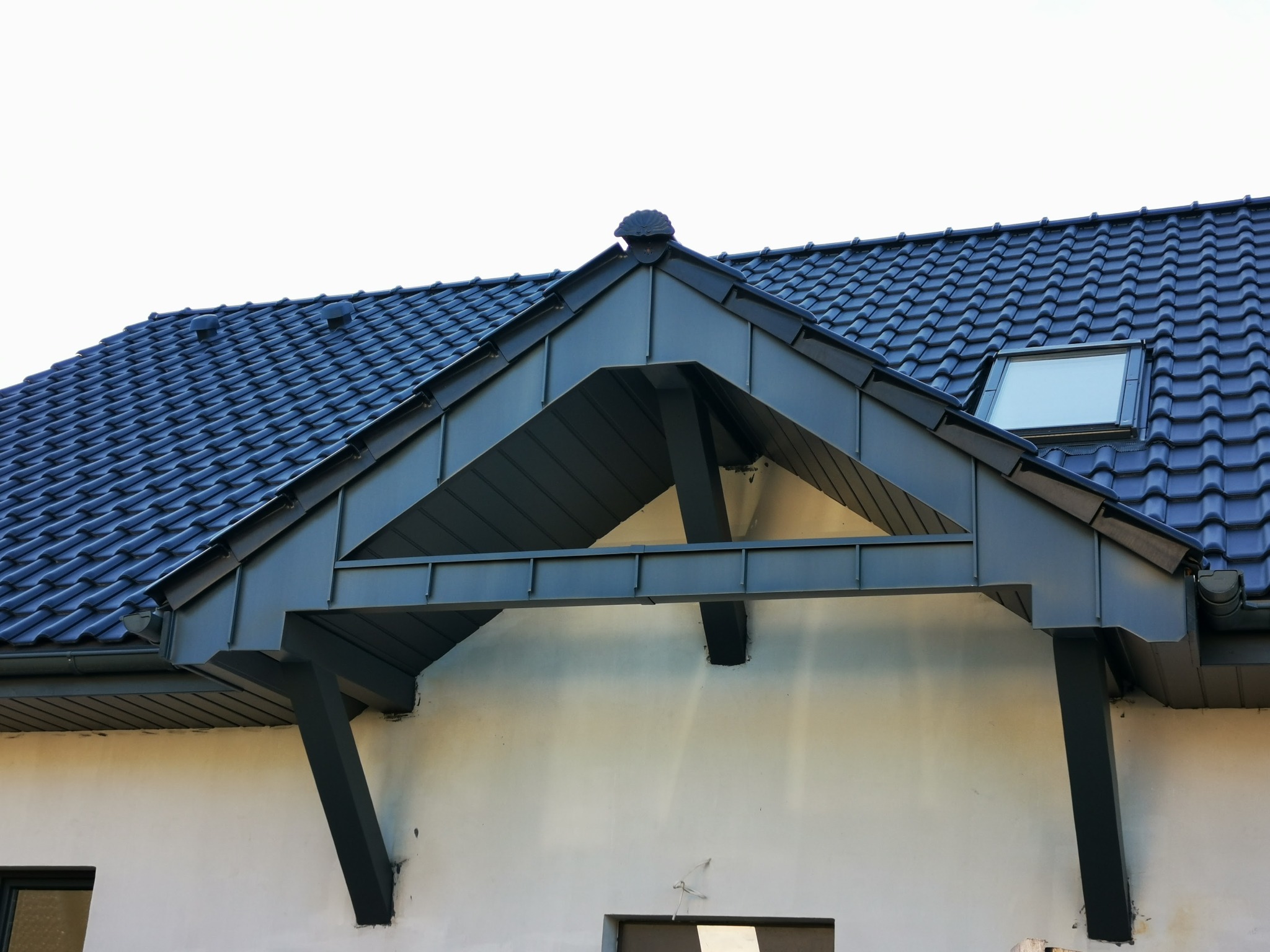Wykończony fragment dachu z ciemnej dachówki ceramicznej, okno dachowe, obróbki blacharskie w kolorze grafitowym, widok na szczyt budynku z elementami konstrukcyjnymi podtrzymującymi zadaszenie.