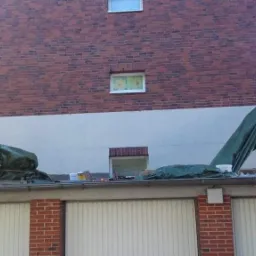 Częściowo otynkowana ceglana ściana budynku z widocznymi rusztowaniami i plandekami ochronnymi, garaże poniżej.