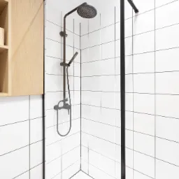 Nowoczesna kabina prysznicowa z czarną armaturą i białymi kwadratowymi płytkami, uzupełniona drewnianą szafką wiszącą.