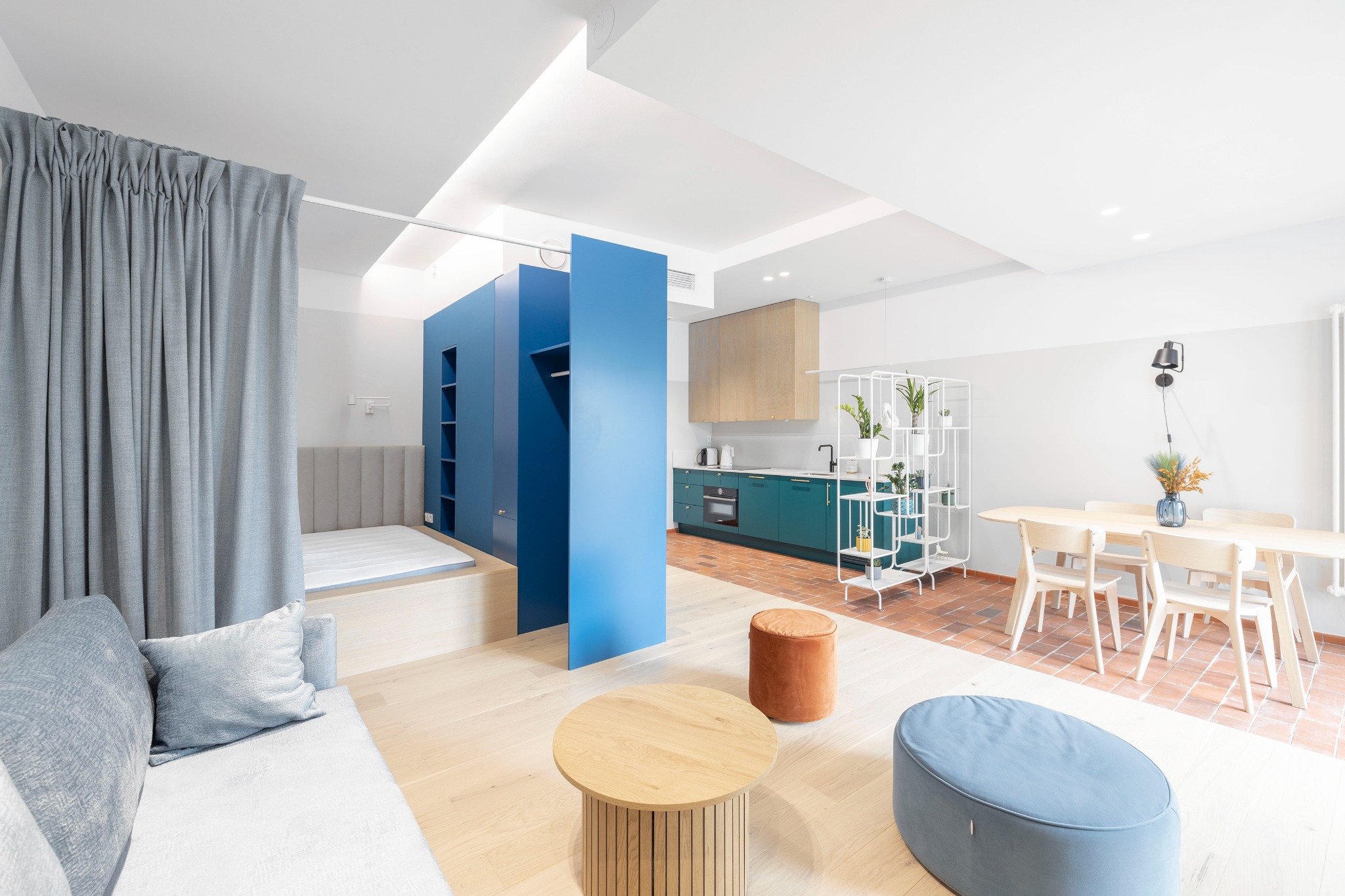 Przestronne wnętrze mieszkania z podziałem na strefy: sypialnia oddzielona zasłoną, niebieska garderoba, kuchnia z zielonymi szafkami i jadalnia z drewnianym stołem na tle ceglanej podłogi.