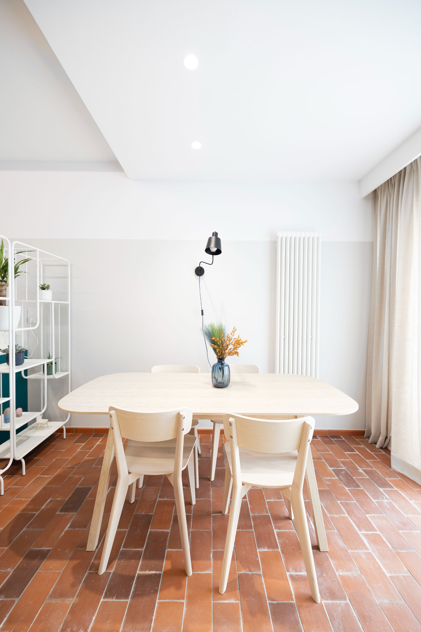 Jasne, minimalistyczne wnętrze jadalni z drewnianym stołem, krzesłami i terakotową podłogą, oświetlone lampą ścienną i naturalnym światłem z okna.