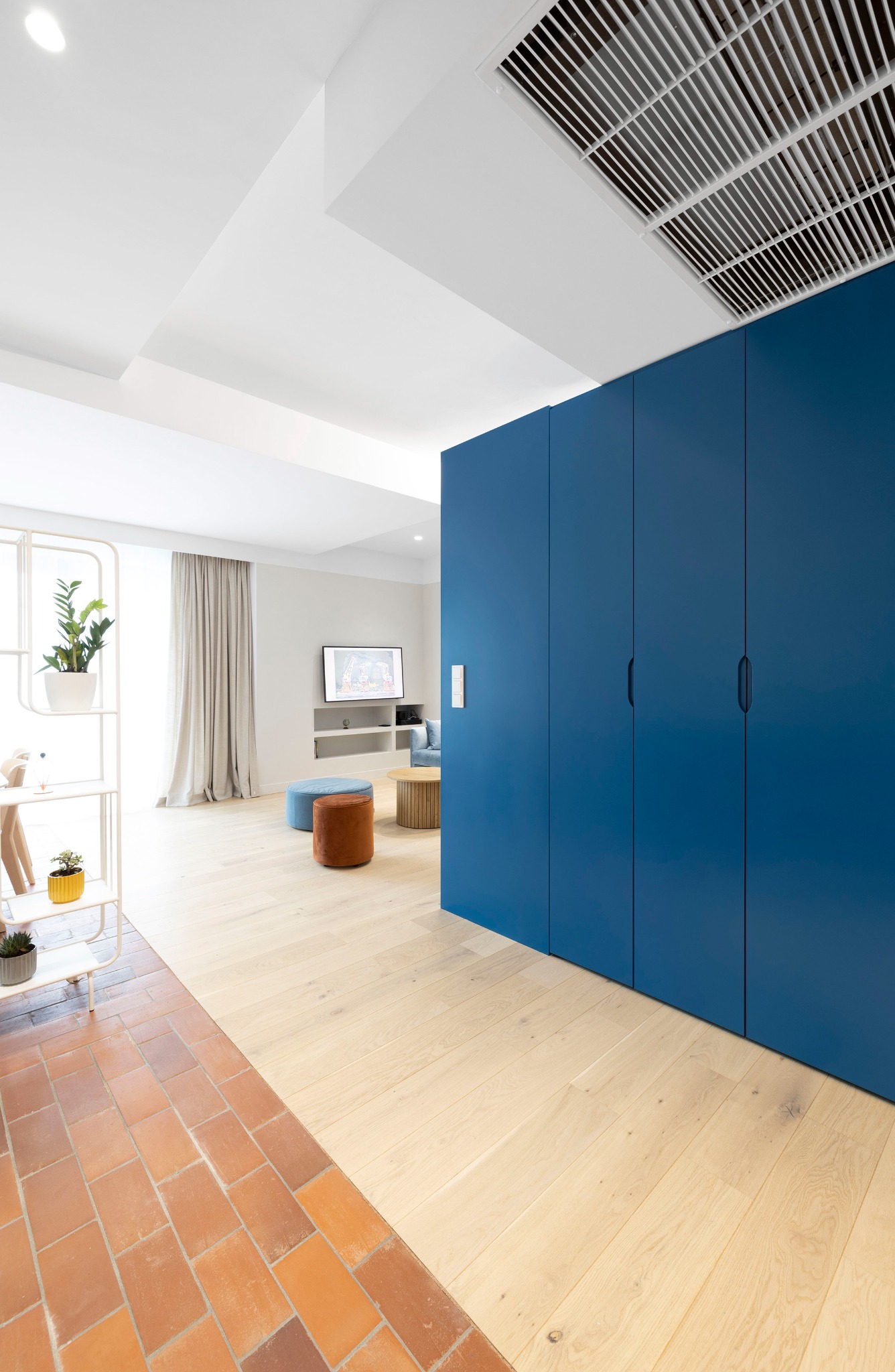 Nowoczesne wnętrze z podziałem podłogi na terakotę i jasne drewno, niebieska zabudowa szafy, widok na salon z pufami i telewizorem.
