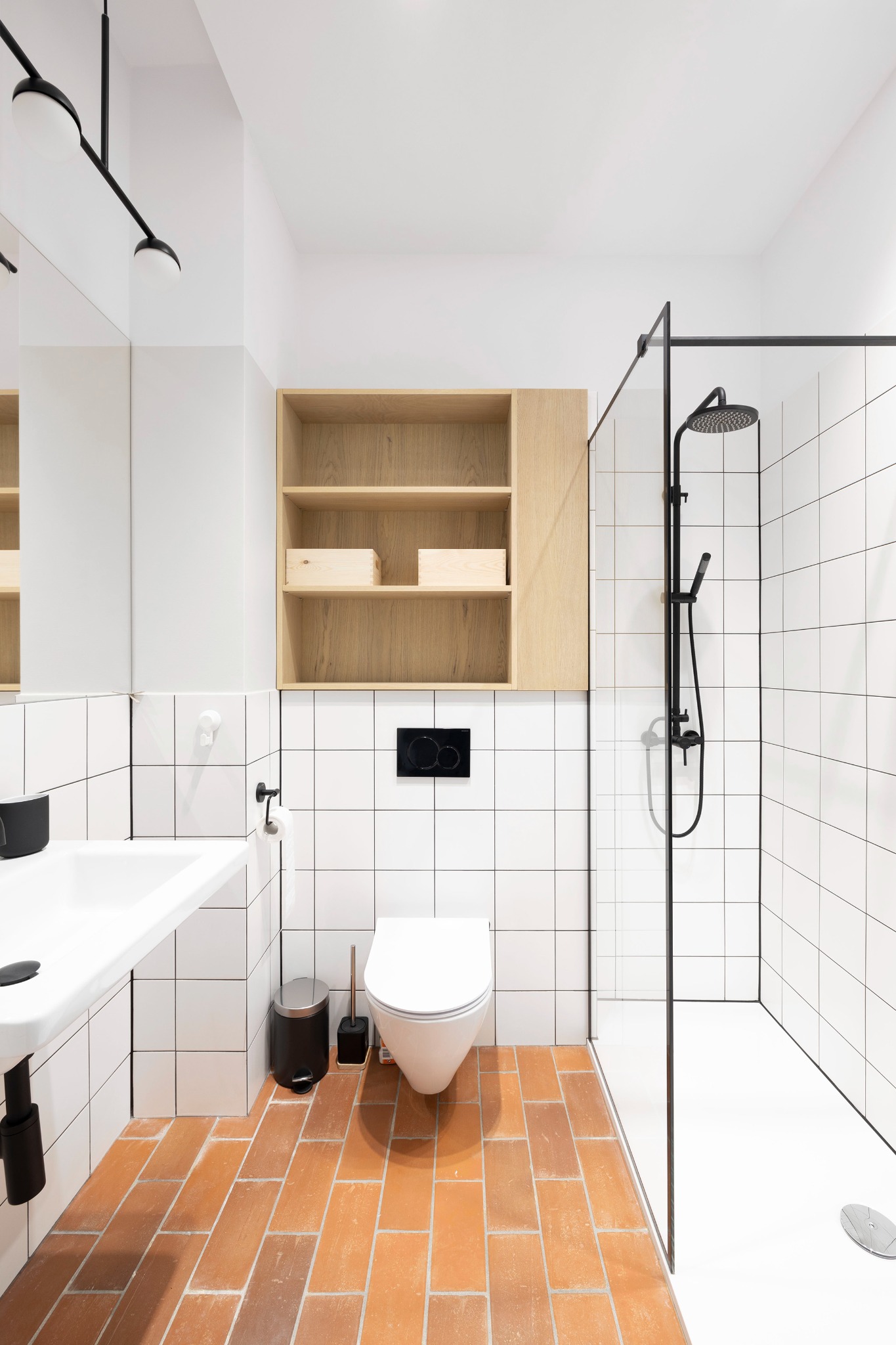 Nowoczesna łazienka z terakotową podłogą, białymi kafelkami, kabiną prysznicową z czarnymi elementami i wiszącą szafką z jasnego drewna.