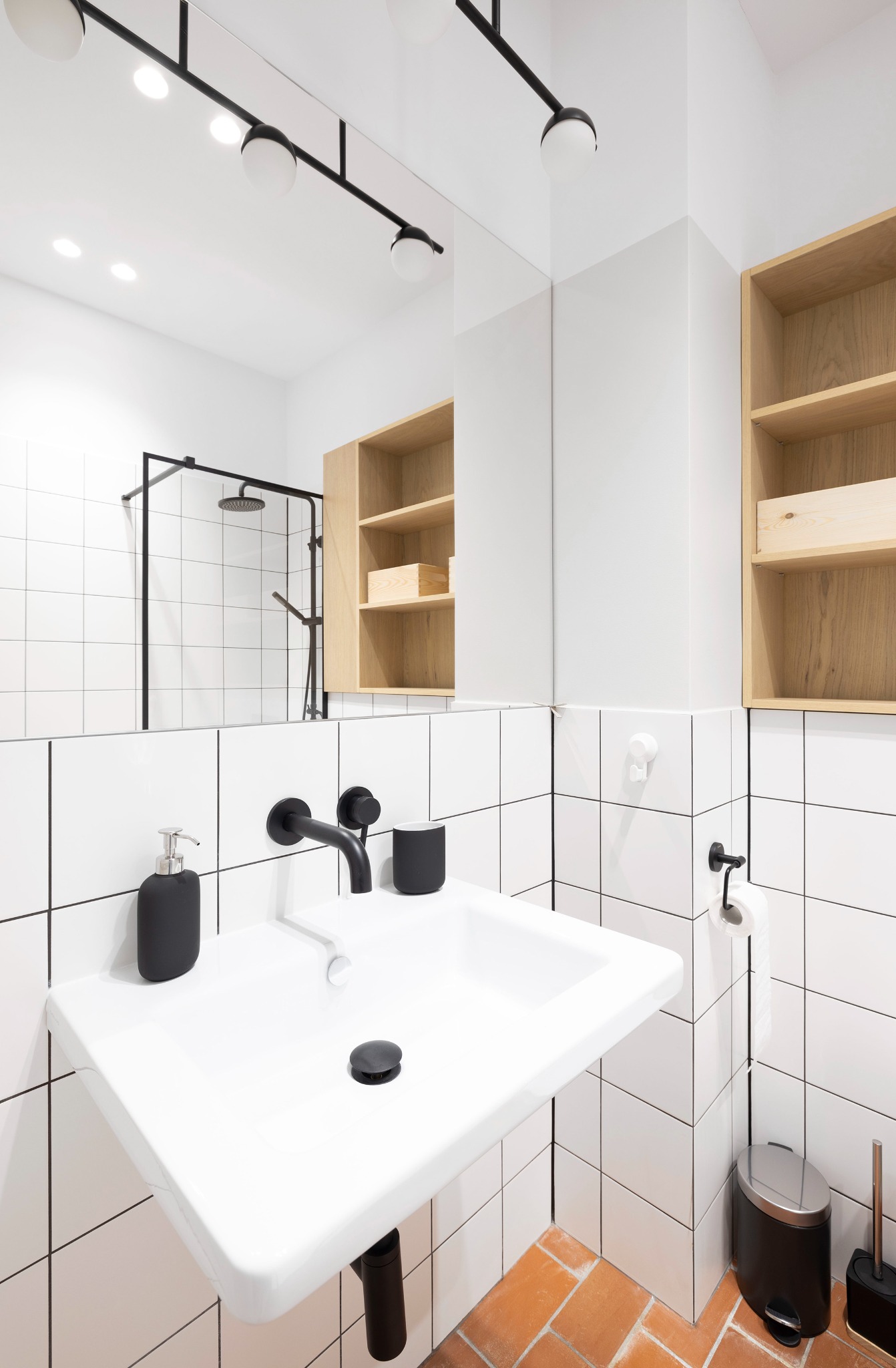 Minimalistyczna łazienka z białą umywalką, czarną armaturą, lustrem odbijającym kabinę prysznicową i drewnianymi półkami, na podłodze terakota.