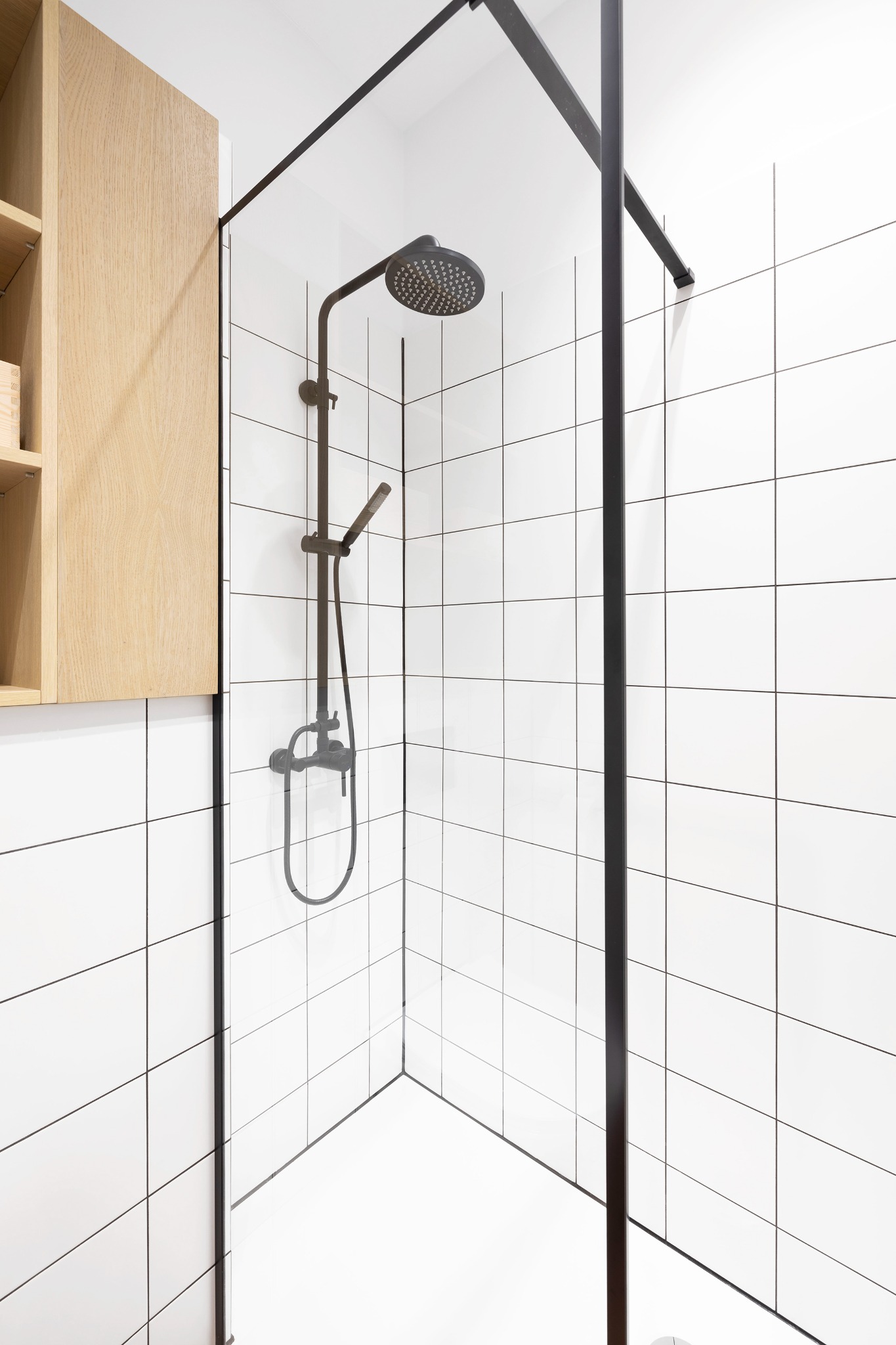 Nowoczesna kabina prysznicowa z czarną armaturą i białymi kwadratowymi płytkami, uzupełniona drewnianą szafką wiszącą.