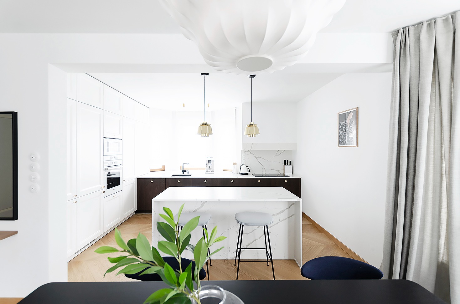 Jasna kuchnia z wyspą, widok z jadalni, parkiet w jodełkę, dekoracyjna lampa sufitowa i minimalistyczne dodatki.