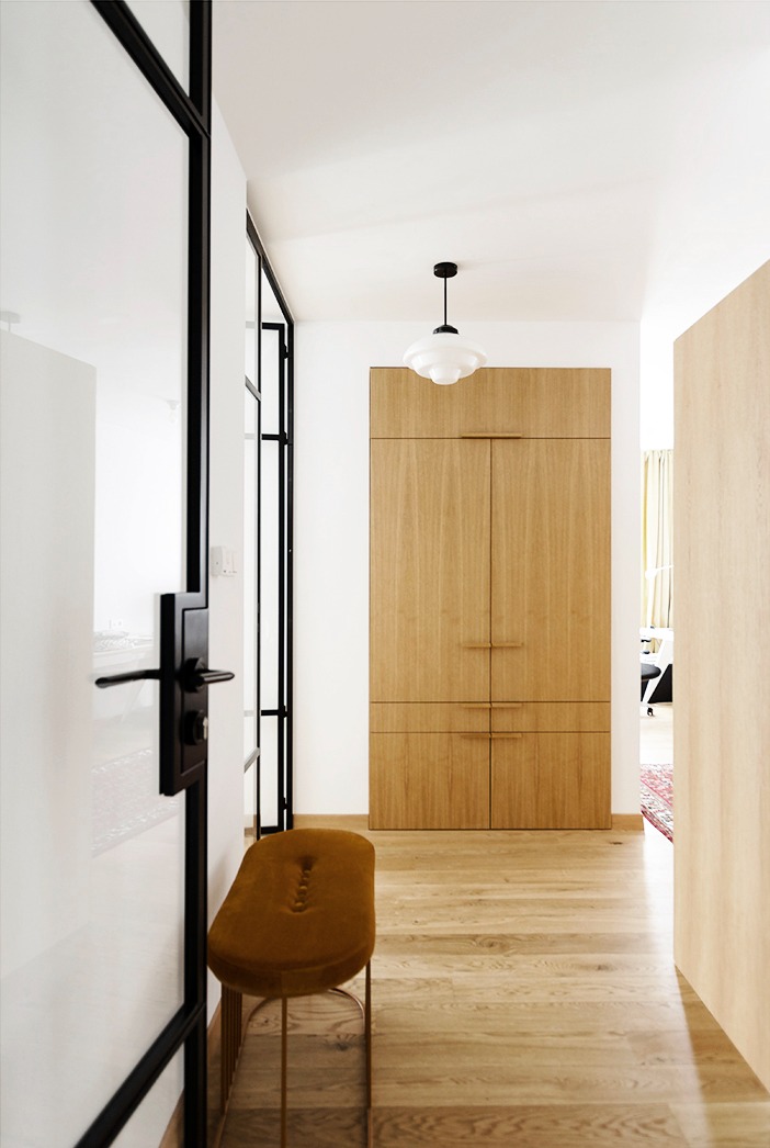 Nowoczesny przedpokój z drewnianą zabudową szafy, czarną ramą szklanych drzwi i musztardowym pufem na złotych nóżkach, oświetlony lampą w stylu retro.