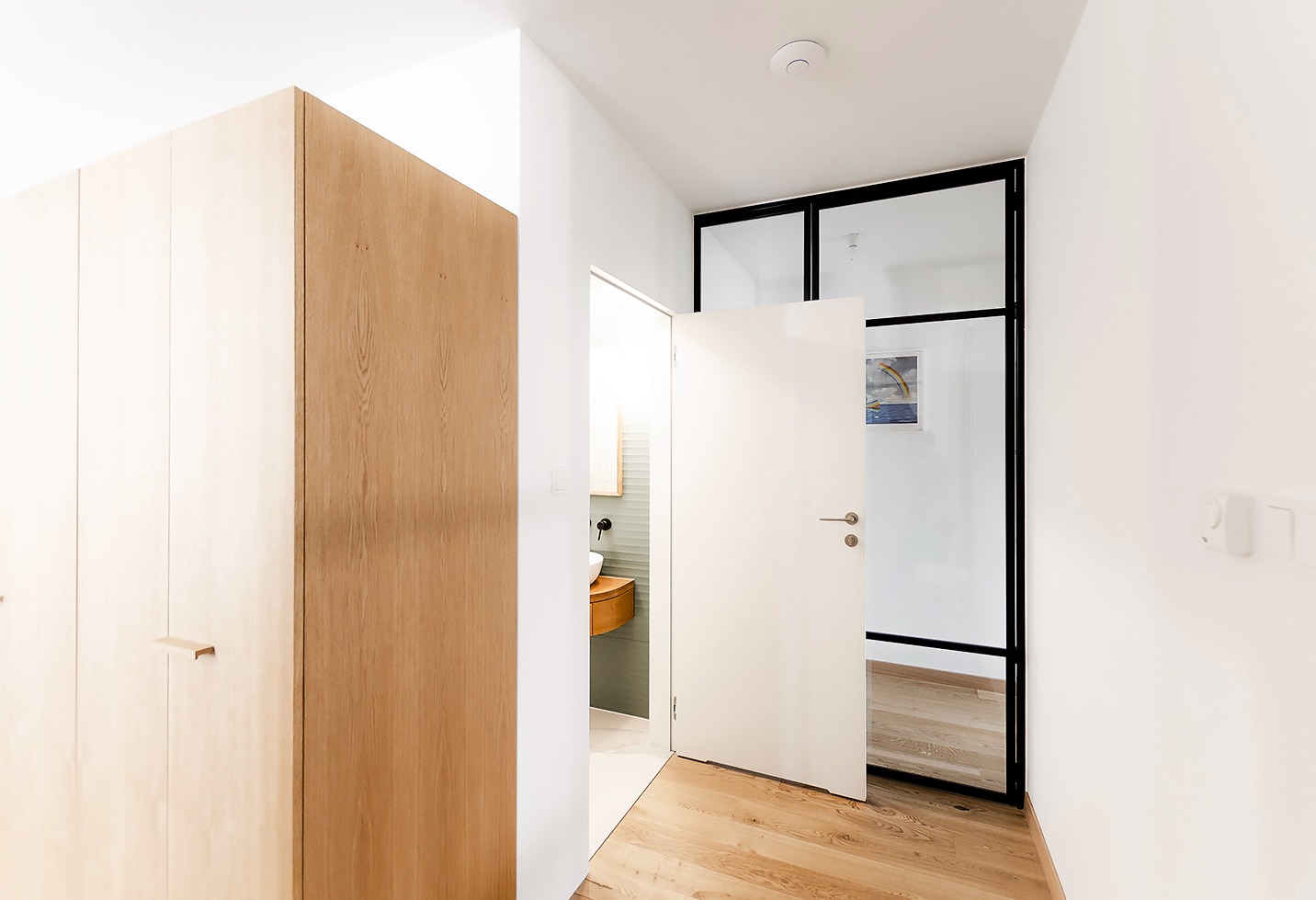 Nowoczesne wnętrze: drewniana szafa w zabudowie, otwarta biała łazienka z drewnianą umywalką oraz przeszklone drzwi z czarnymi ramami, parkiet z jasnego drewna.