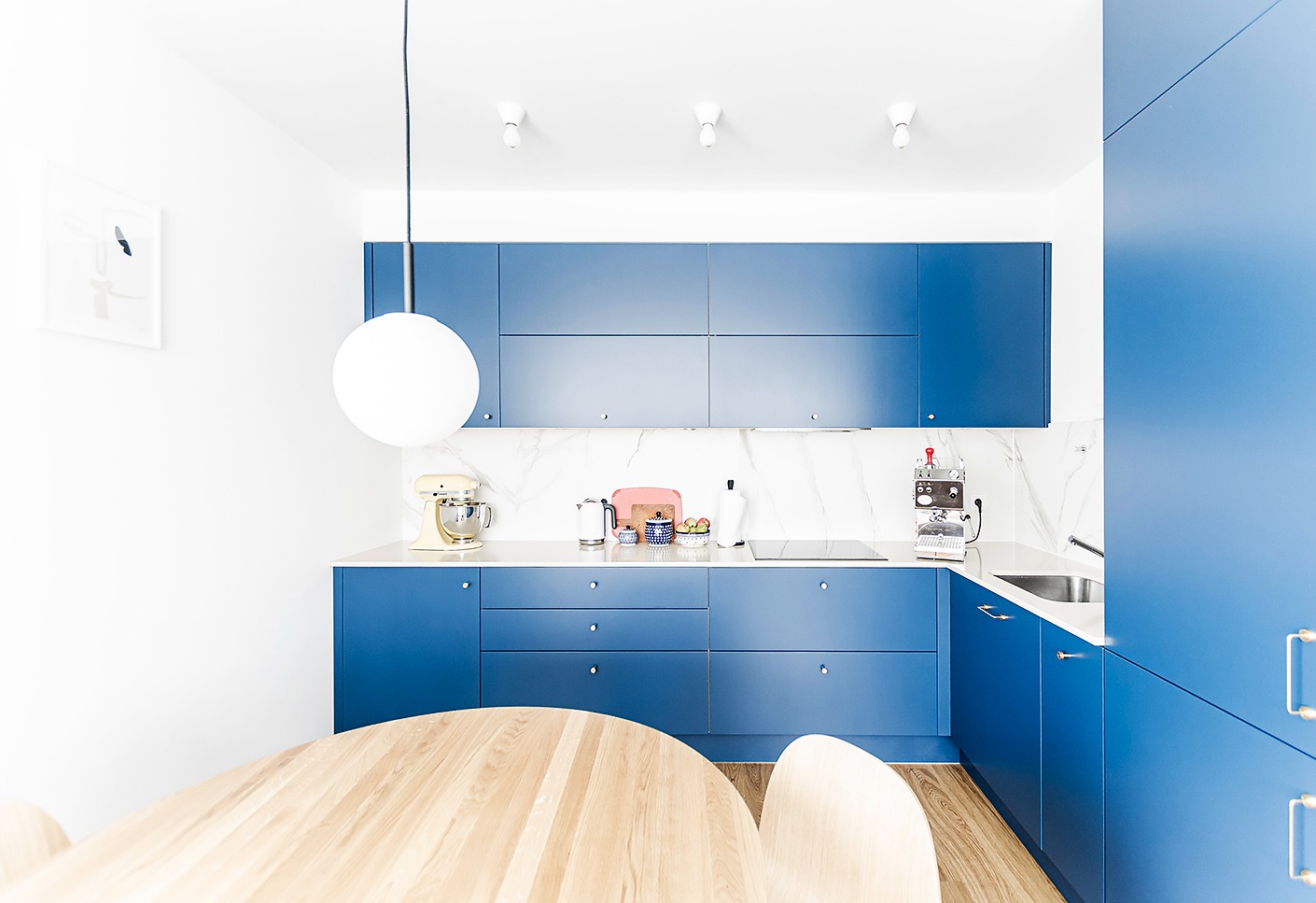 Nowoczesna kuchnia z niebieskimi szafkami i białym blatem z marmurowym wzorem, okrągły drewniany stół i biała lampa kula.