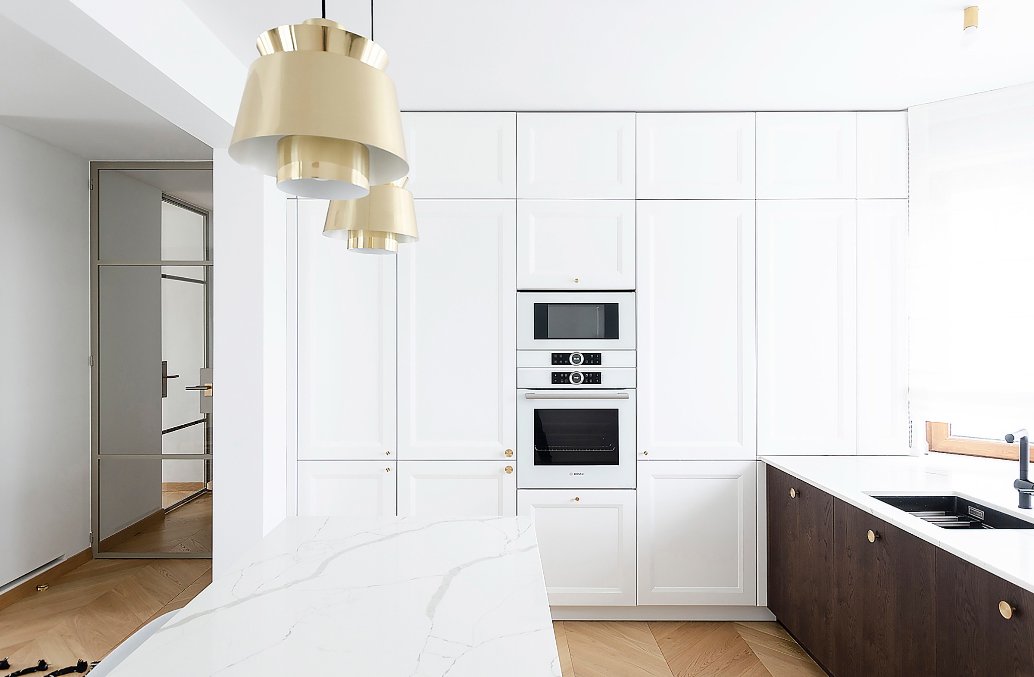 Jasna kuchnia z białymi szafkami, sprzętem AGD w zabudowie i marmurowym blatem wyspy, oświetlona złotymi lampami wiszącymi.