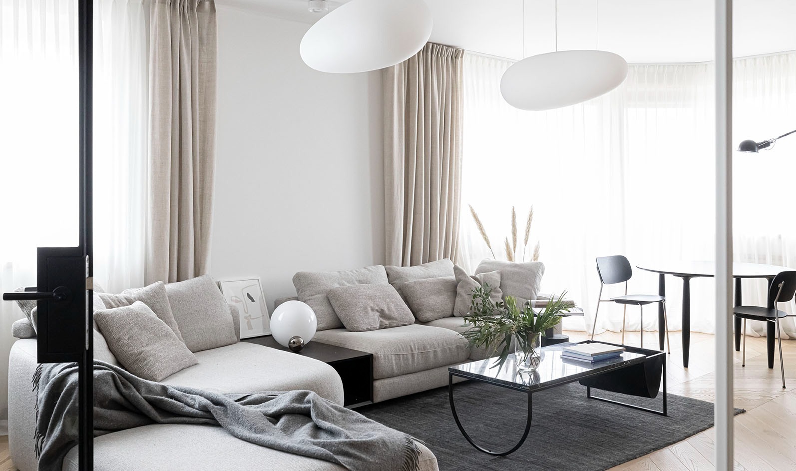 Jasny salon z szarą sofą narożną, szklanym stolikiem kawowym z czarną ramą, owalnymi lampami wiszącymi, czarnymi krzesłami i okrągłym stołem w tle, oraz zasłonami w odcieniach beżu i bieli.