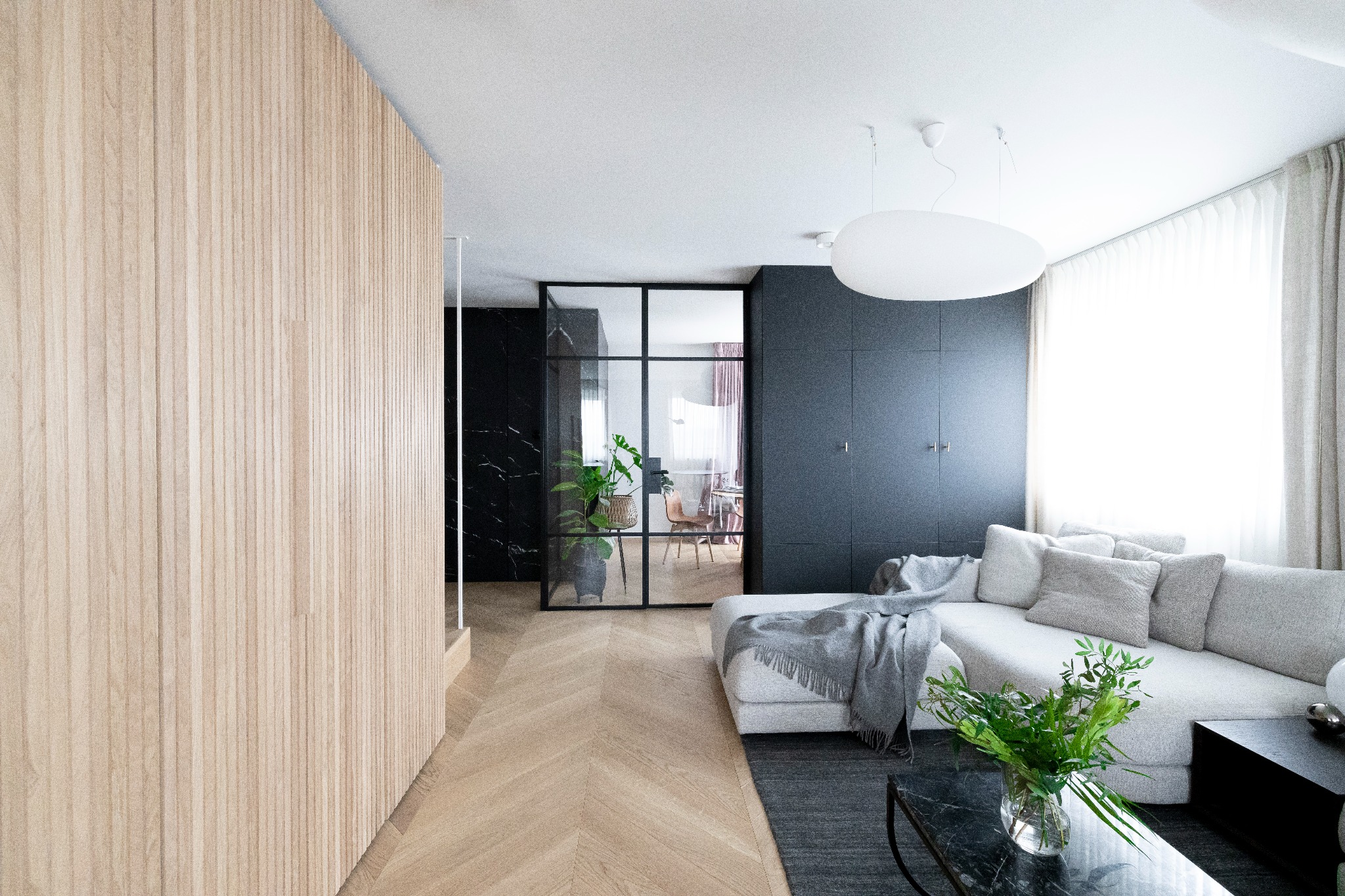 Jasne, przestronne wnętrze z drewnianą ścianą, parkietem w jodełkę, szklaną ścianą działową i narożną sofą z szarym pledem. W tle widoczna marmurowa ściana i minimalistyczne oświetlenie.
