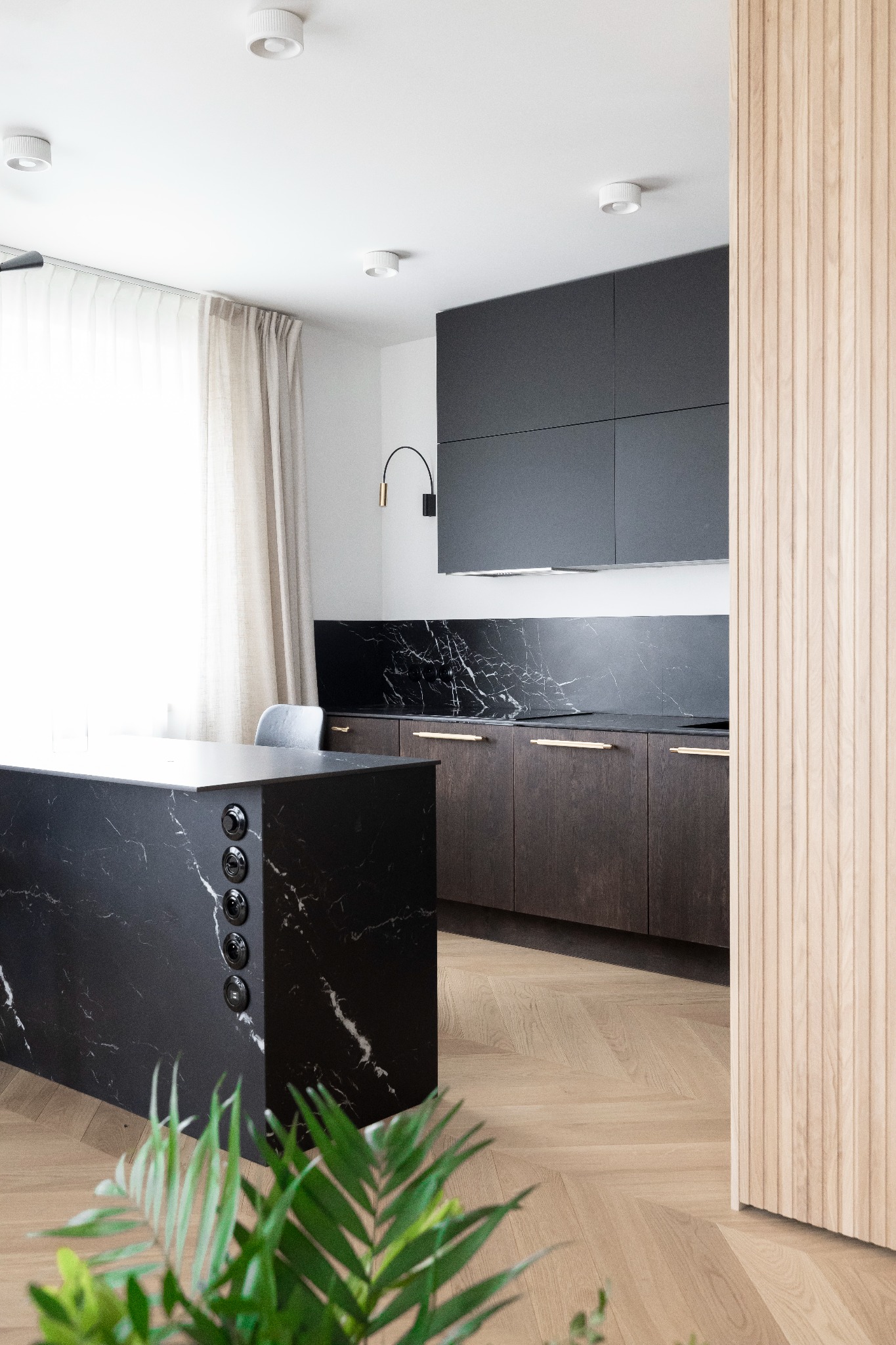 Nowoczesna kuchnia z wyspą z czarnego marmuru, drewnianymi szafkami i parkietem w jodełkę, oświetlona punktowymi lampami sufitowymi i designerskim kinkietem.