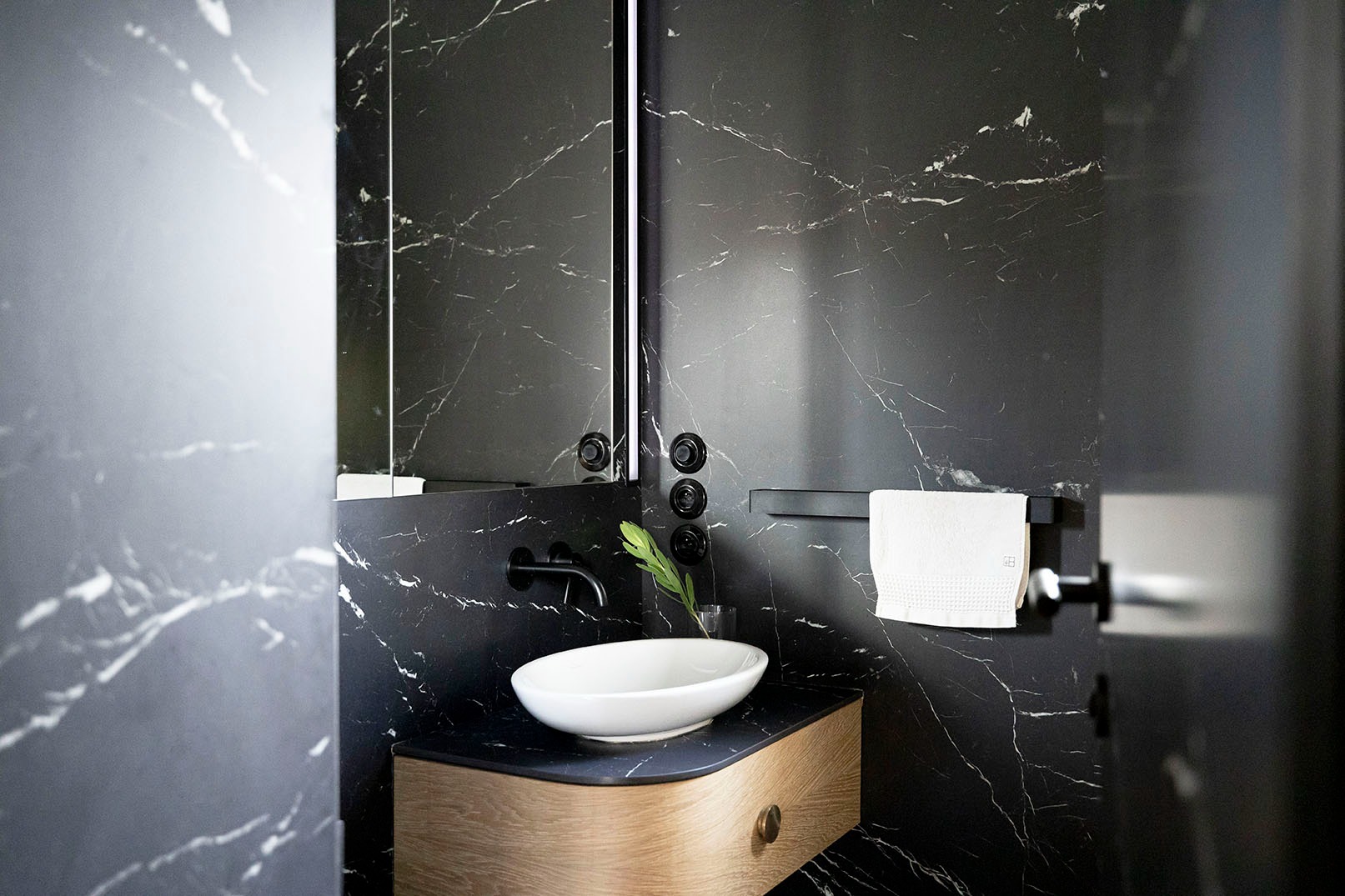 Nowoczesna łazienka z czarnymi marmurowymi ścianami, owalną białą umywalką na drewnianej szafce i minimalistyczną armaturą.