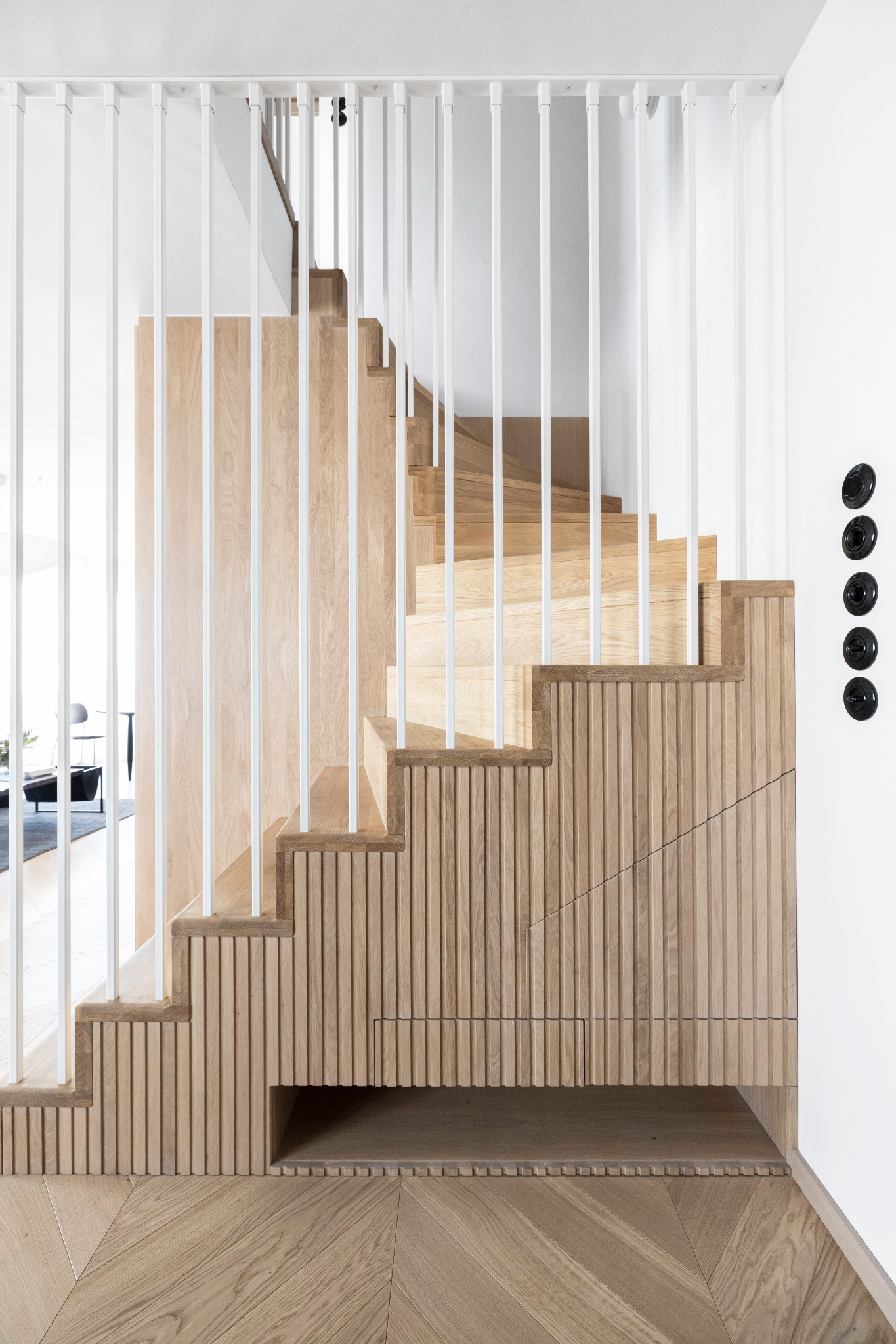 Drewniane schody z białymi, pionowymi balustradami, wykończone panelami z rowkowanego drewna i podświetloną wnęką, na parkiecie ułożonym w jodełkę.