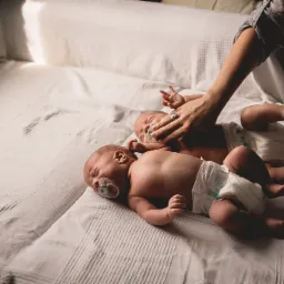 Dwoje noworodków w pieluchach leży na białym łóżku, jedno z nich ma smoczek w ustach, a dłoń osoby dorosłej z pierścionkiem na palcu delikatnie dotyka drugiego dziecka.