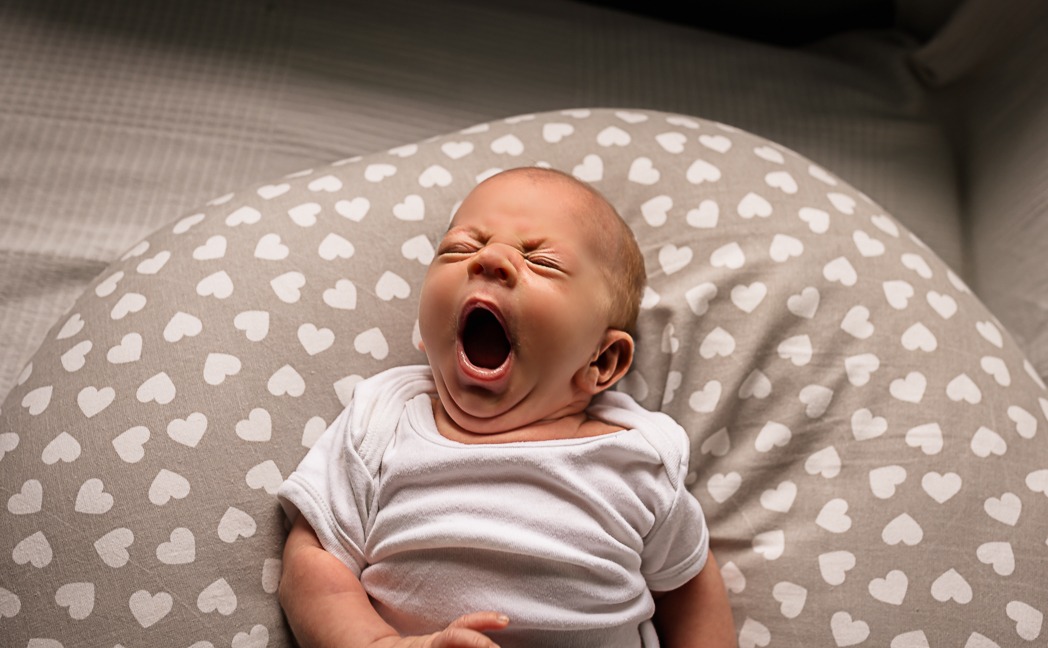 Noworodek w białym body ziewa, leżąc na poduszce w szare serduszka.