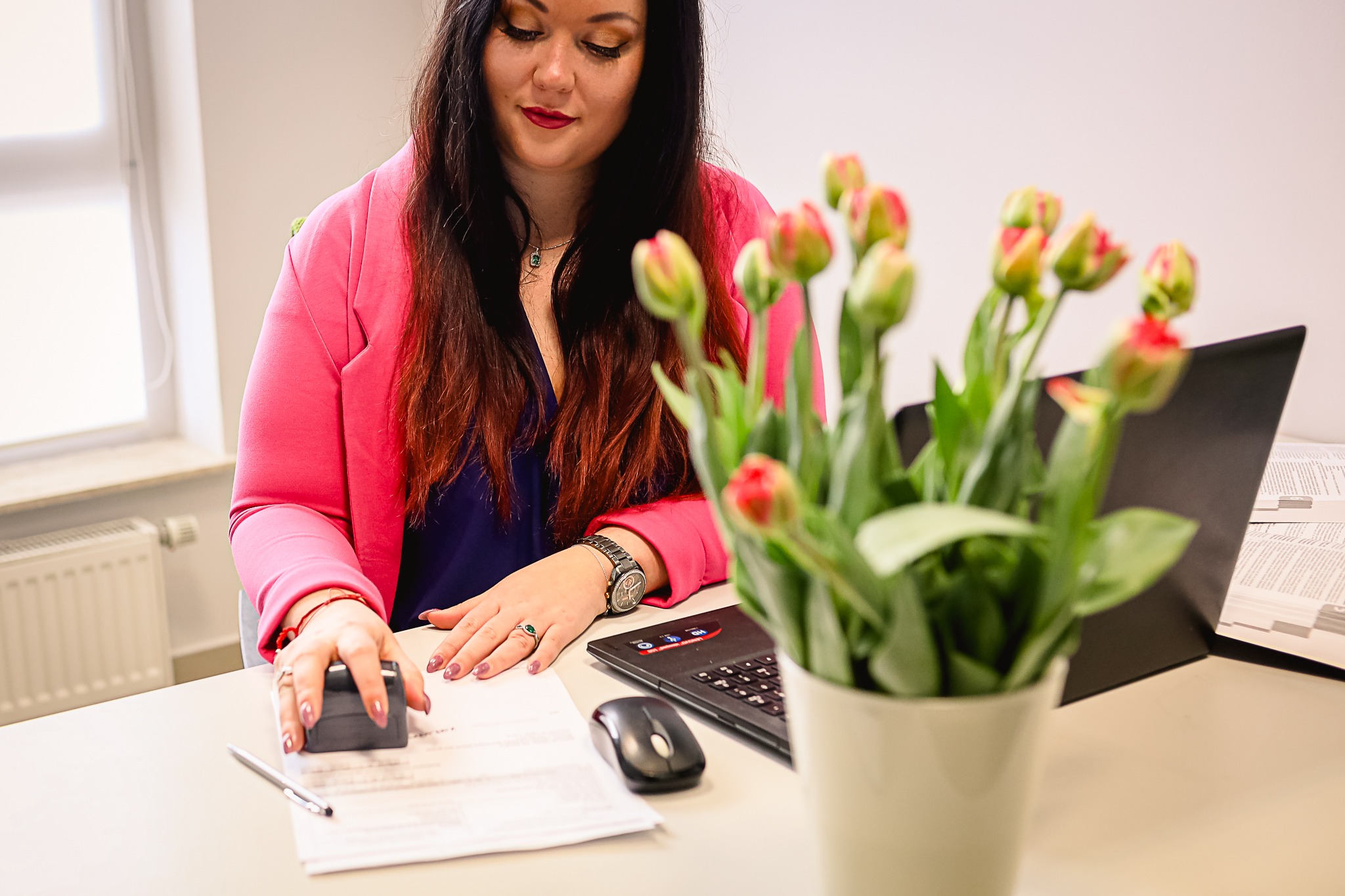 Kobieta w różowej marynarce przy biurku stempluje dokument, obok laptop, myszka, długopis i bukiet tulipanów w wazonie.
