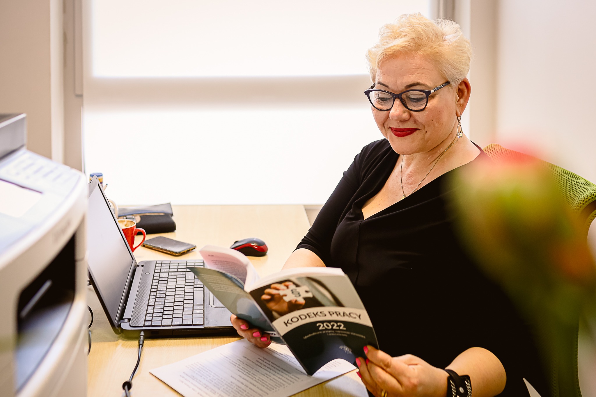 Kobieta w okularach czyta Kodeks Pracy 2022 przy biurku z laptopem, drukarką i czerwonym kubkiem.