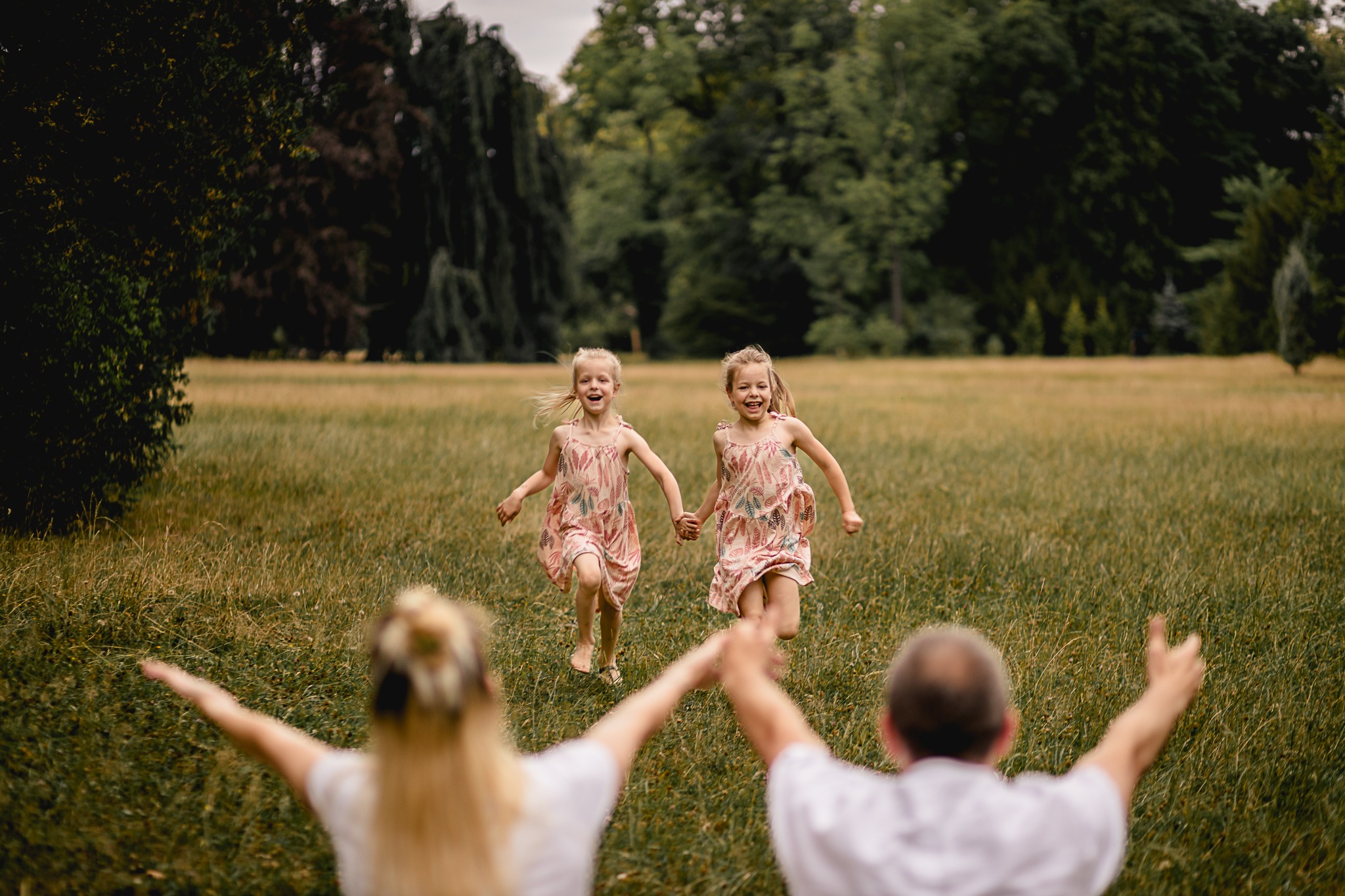 Dwie uśmiechnięte dziewczynki w zwiewnych sukienkach biegną trzymając się za ręce w kierunku rozradowanych rodziców na łące w parku.