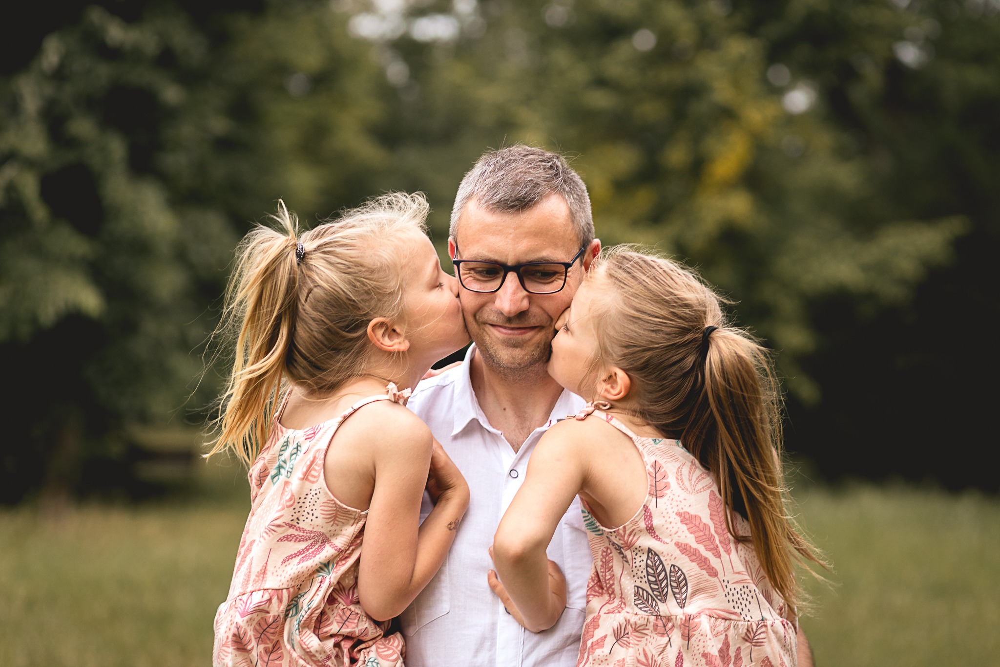 Uśmiechnięty tata w okularach całowany w policzki przez dwie blond córeczki w sukienkach w plenerze.
