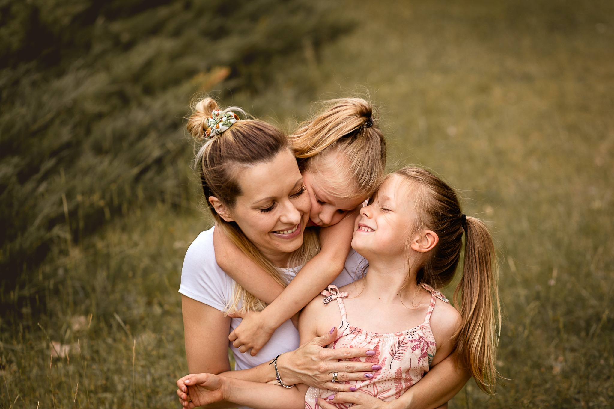 Uśmiechnięta mama tuląca dwie córki na tle zieleni, z włosami spiętymi w koki z ozdobnymi gumkami.
