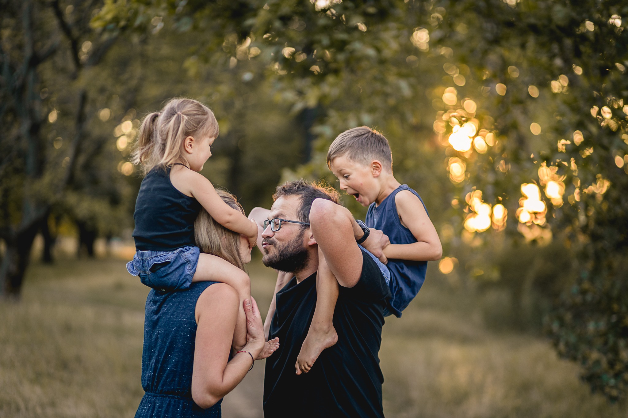 Rodzina w plenerze: mama i tata niosą na ramionach córkę i syna, chłopiec z otwartymi ustami, jasne bokeh w tle.