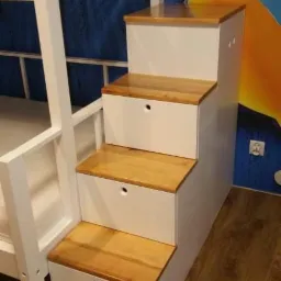 Białe łóżko piętrowe z drewnianymi schodami-szufladami, widoczne na tle ściany z dziecięcym motywem.