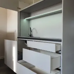 Nowoczesna zabudowa kuchenna w odcieniach szarości i bieli z czarną armaturą, podświetlona listwą LED, z otwartymi szufladami i uchylnymi drzwiczkami szafki.