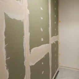 Ściana z płyt gipsowo-kartonowych w trakcie prac, widoczne łączenia płyt wypełnione szpachlą, wstępnie przygotowane do dalszej obróbki, z widocznymi punktowymi wgłębieniami na powierzchni płyt.