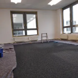 Pokój w trakcie przygotowania do malowania: zabezpieczona podłoga, okna i grzejniki oklejone taśmą malarską, drabina w centrum, widoczne wiaderko z farbą.