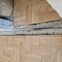Olech Damian - Ułożona parkietowa podłoga w jodełkę, widoczne niedokończone krawędzie przy ścianie z odsłoniętym betonem i instalacjami, worki z materiałami budowlanymi w rogu.
