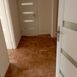 Olech Damian - Parkiet ułożony w jodełkę w jasnym korytarzu z białymi drzwiami z przeszkleniami.