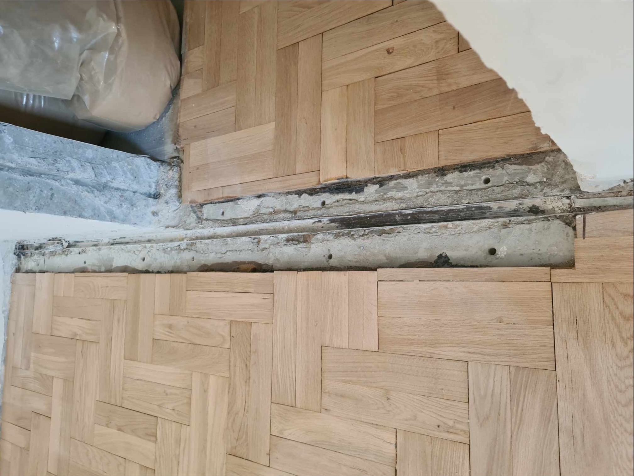 Ułożona parkietowa podłoga w jodełkę, widoczne niedokończone krawędzie przy ścianie z odsłoniętym betonem i instalacjami, worki z materiałami budowlanymi w rogu.