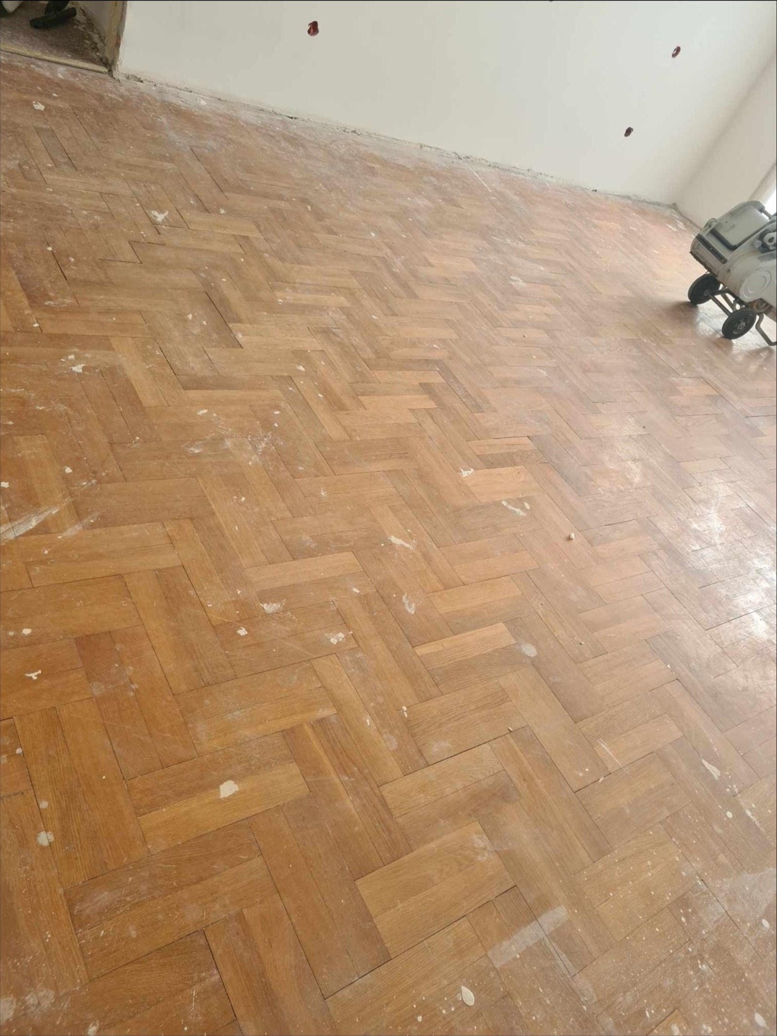 Parkiet ułożony w jodełkę po wstępnym szlifowaniu, widoczne ślady pyłu i maszyna do obróbki drewna w rogu pomieszczenia.