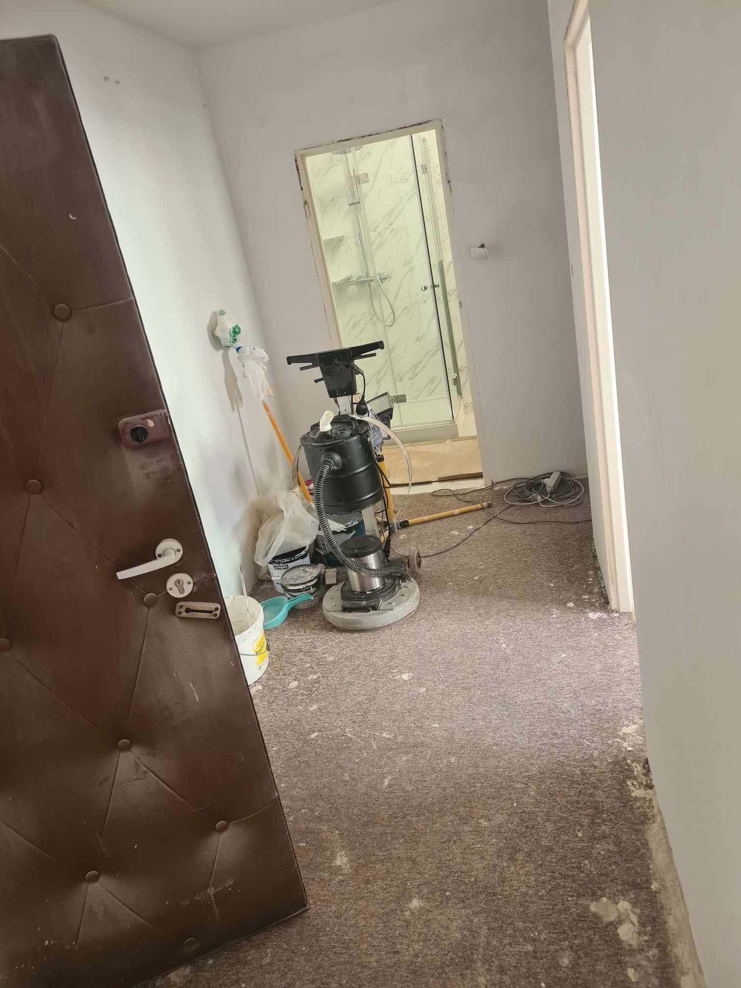 Pokój w trakcie renowacji podłogi, widoczna maszyna do szlifowania parkietu, brudna brązowa podłoga i otwarte drzwi do łazienki z kabiną prysznicową w tle.