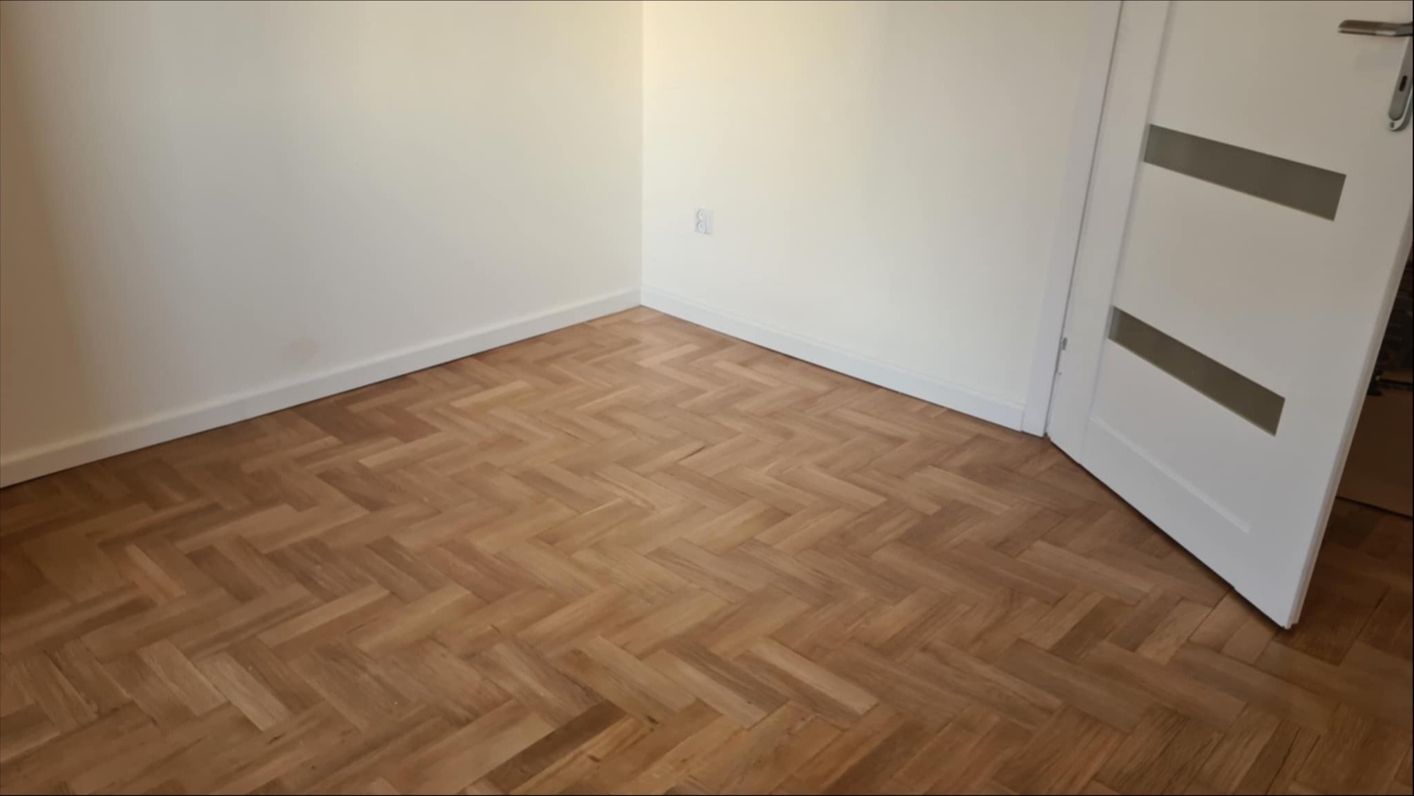 Jasny parkiet ułożony w jodełkę w rogu pokoju z białymi ścianami i otwartymi białymi drzwiami z szarymi paskami.