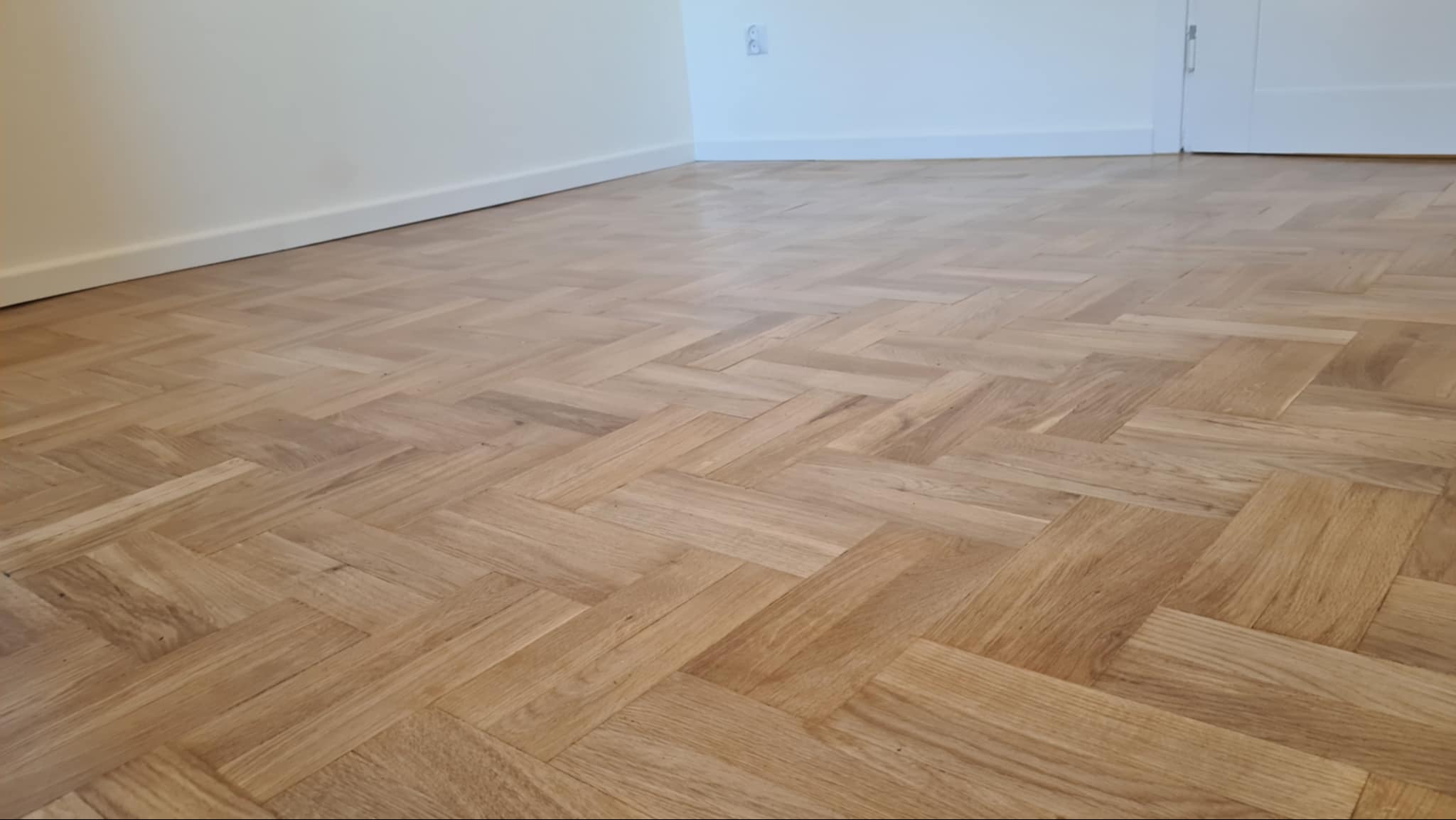 Parkiet ułożony w jodełkę po renowacji, widoczne subtelne refleksy światła na gładkiej powierzchni, ściana w tle pomalowana na jasny odcień beżu.