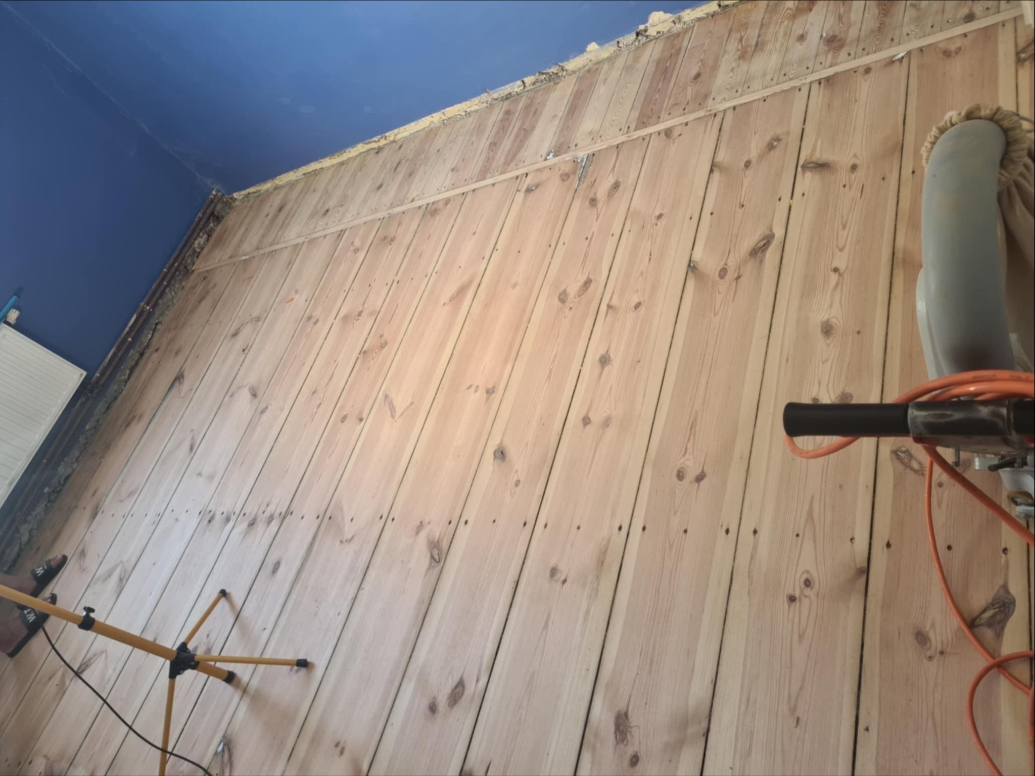 Świeżo wyszlifowana drewniana podłoga z widocznymi śladami po gwoździach, fragment maszyny do szlifowania i żółty statyw lampy oświetleniowej w rogu pomieszczenia z niebieskim sufitem.