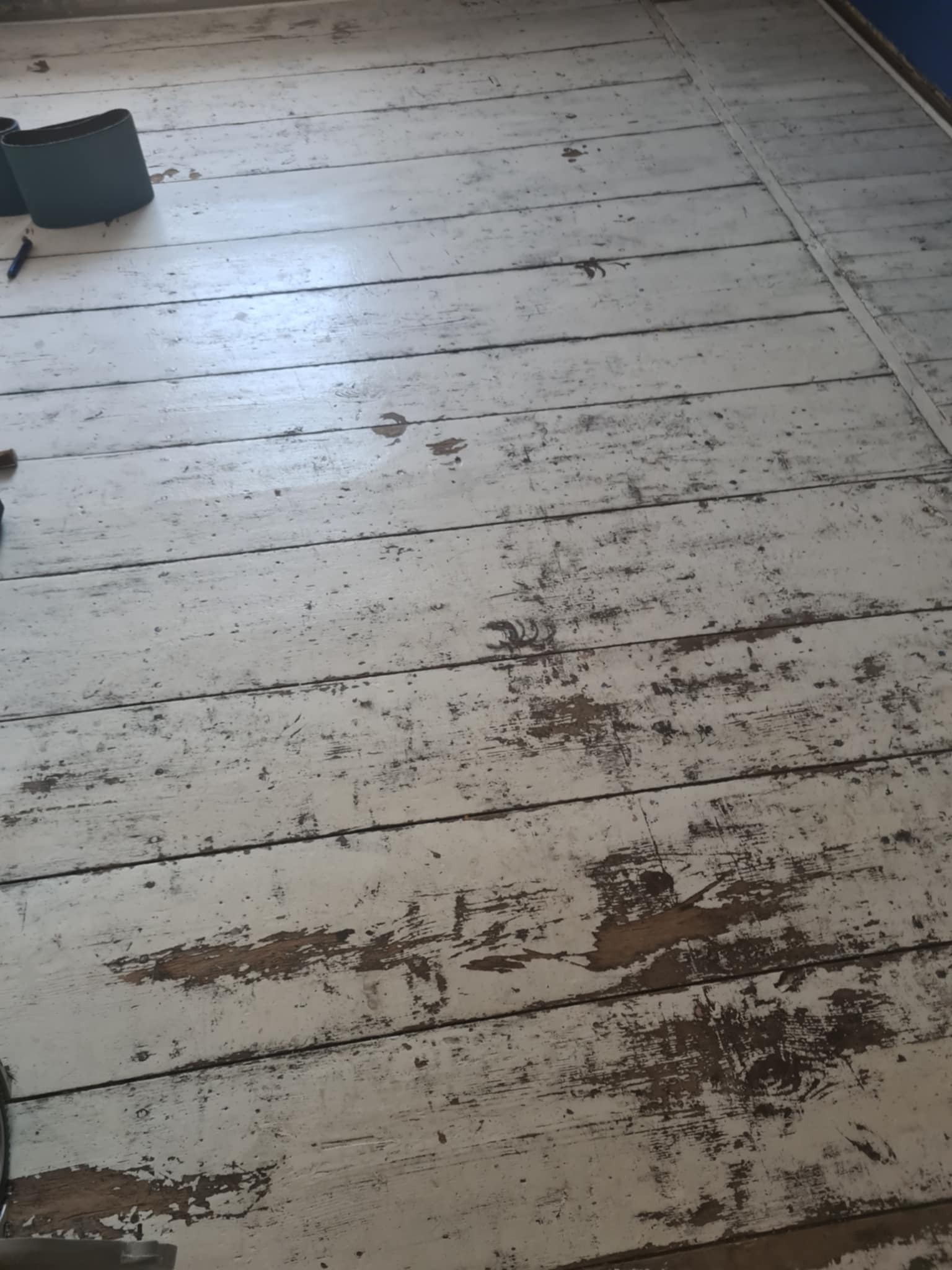 Podłoga z desek, częściowo zeszlifowana z białej farby, widoczne ślady po szlifowaniu, obok leżą pasy ścierne.