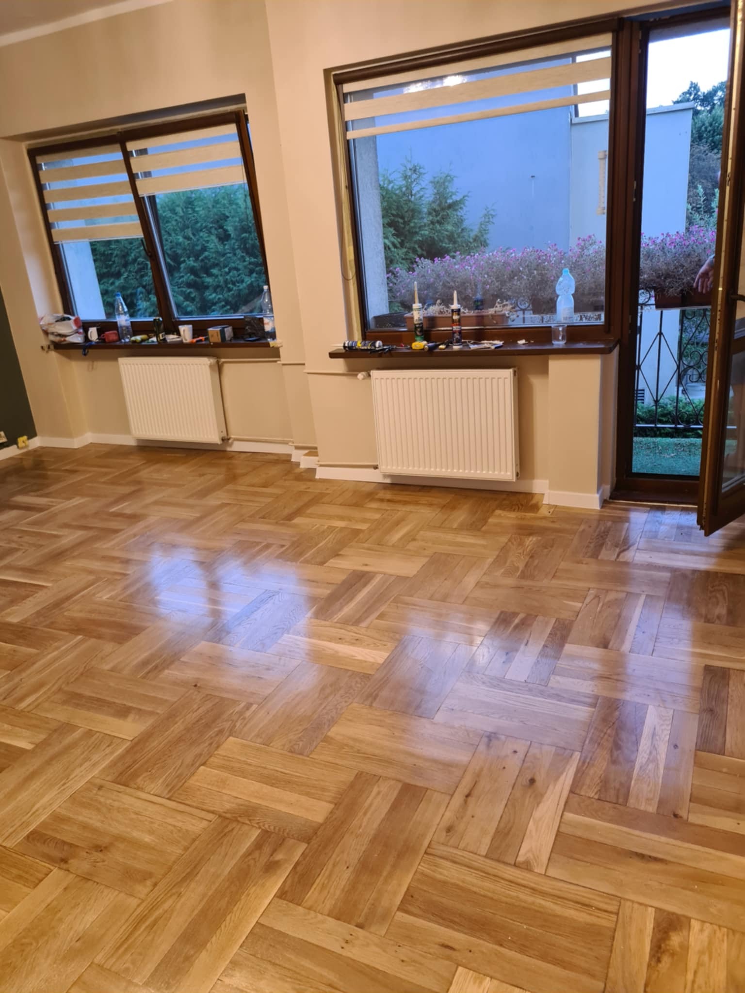 Jasny parkiet ułożony we wzór jodełki francuskiej w słonecznym salonie z dwoma oknami i otwartymi drzwiami balkonowymi.